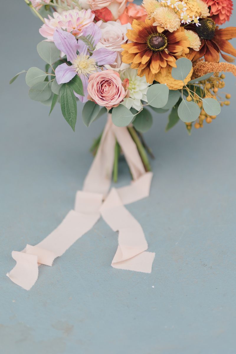 Colorful wedding bouquet with sunflowers, roses, and lavender blooms tied with a cream ribbon on a blue surface.
