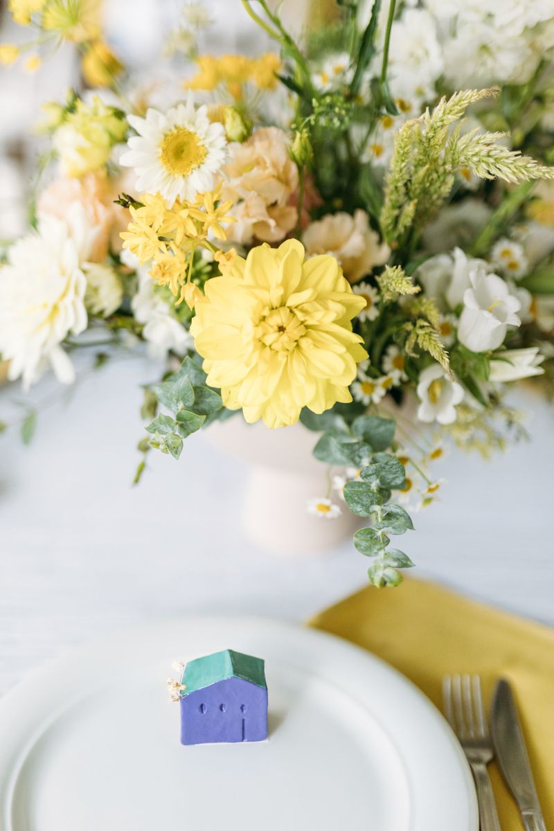 Elegant table setting with yellow dahlia floral centerpiece and small blue house figurine on white plate.
