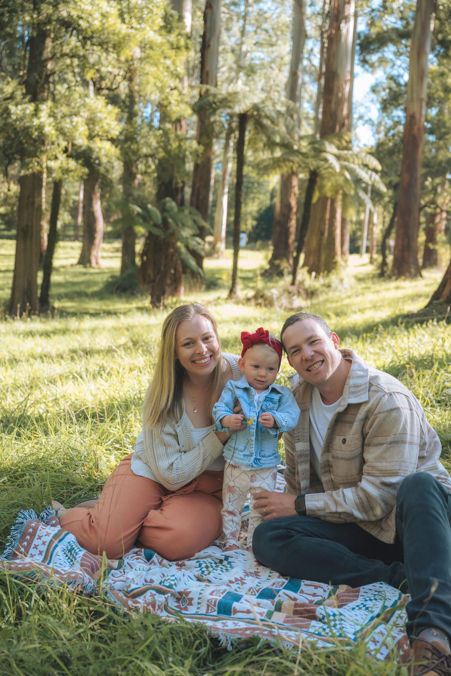 Family of three smiling on a colorful blanket in a sunlit forest park, with parents and baby girl in a red bow.