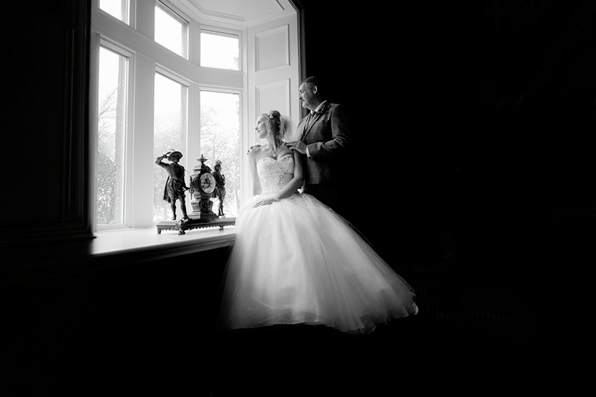 Black and white wedding photo of bride and groom silhouetted by a large bay window in an elegant room.