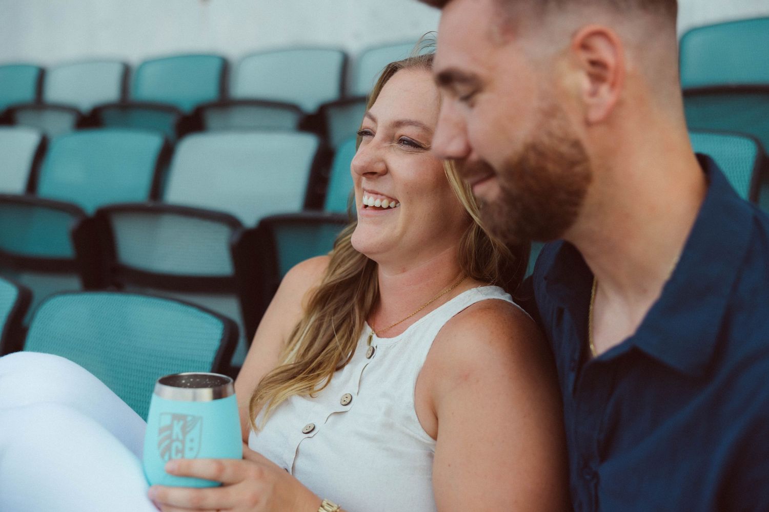 A closeup of a newly engaged couple sitting and talking in teal stadium seats following their surprise proposal at CPKC stadium in KCMO.