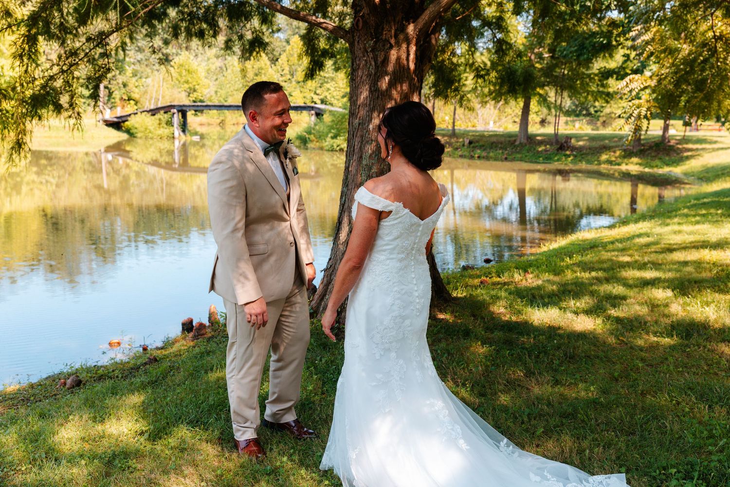 Bride and groom share a romantic first look moment by a serene pond surrounded by lush green trees.
