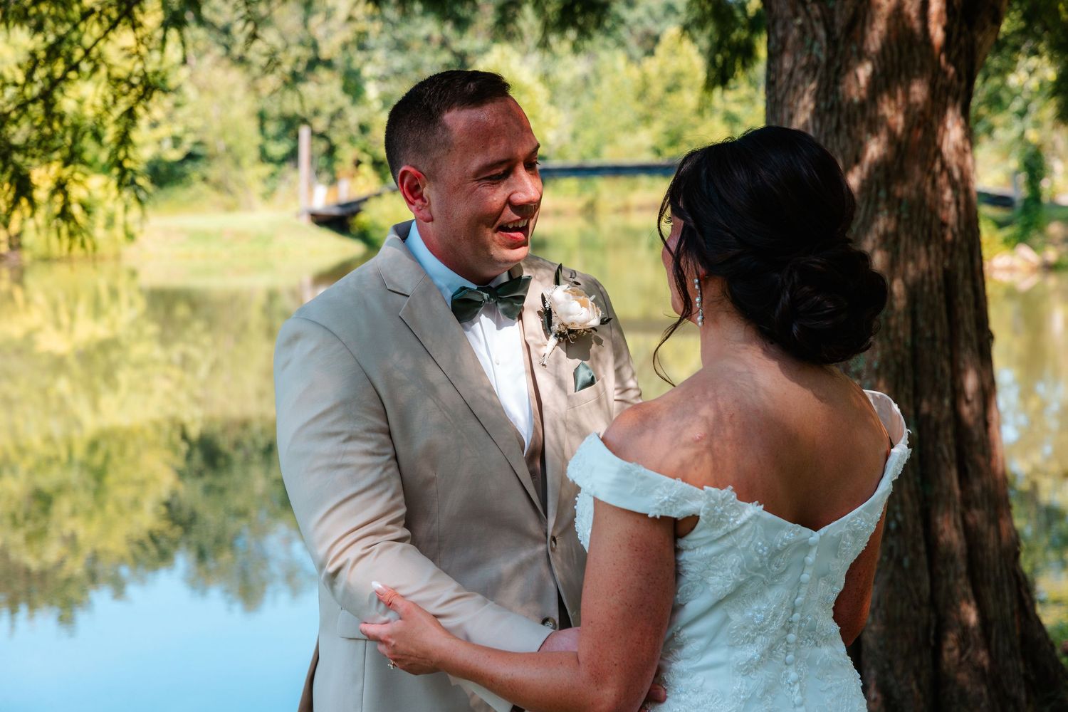 Groom smiles emotionally as bride touches his face during outdoor wedding first look near a pond.