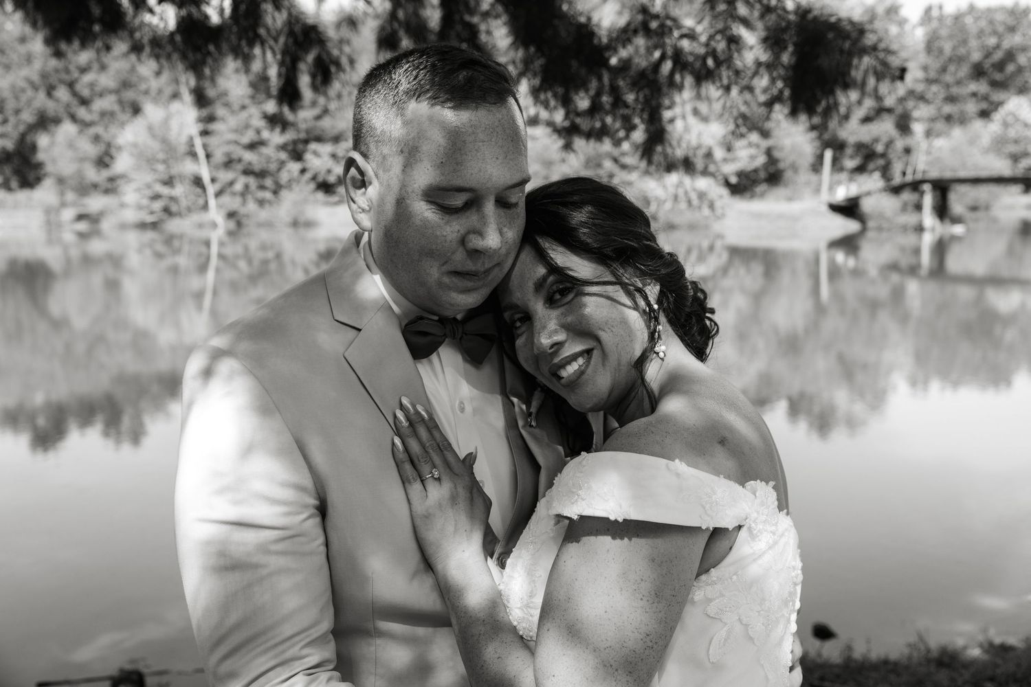 Black and white wedding photo of bride and groom embracing tenderly by a serene lake surrounded by lush trees.