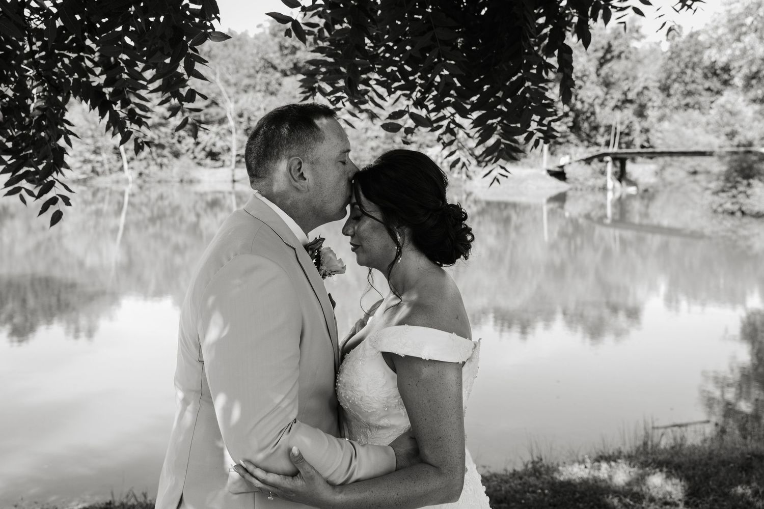 Black and white photo of a couple embracing and kissing near a reflective pond surrounded by lush trees on their wedding day.