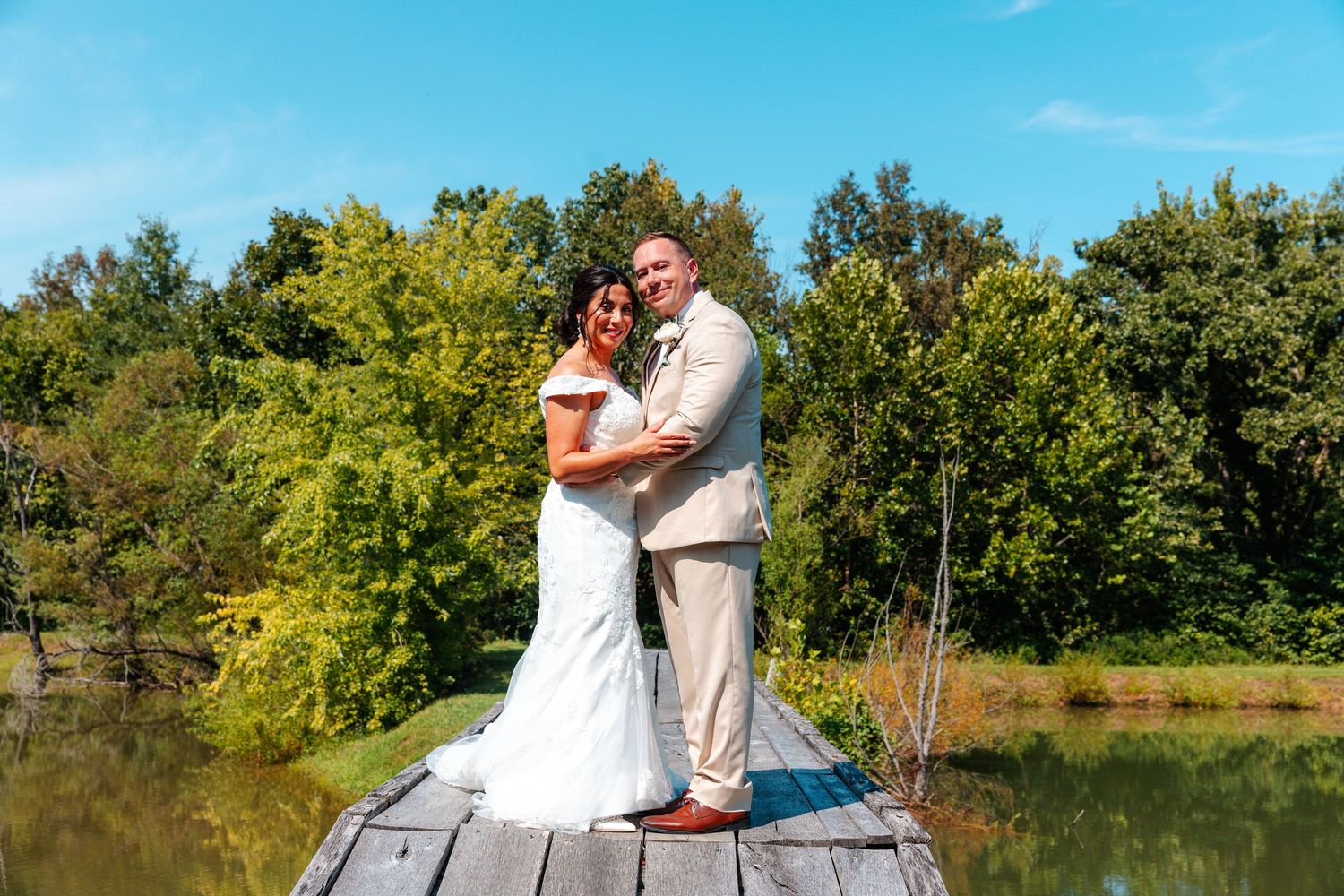 Bride and groom posing on a wooden dock by a lake surrounded by lush green trees on a sunny wedding day.