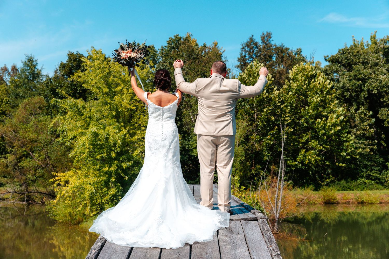 Bride and groom celebrate on a wooden dock overlooking a serene lake surrounded by lush green trees on their wedding day.