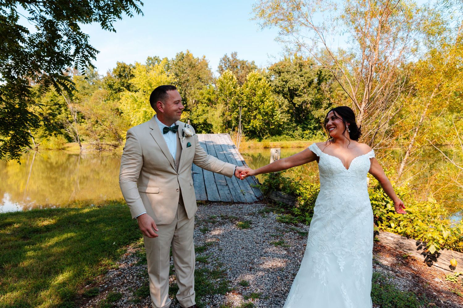 Bride and groom hold hands outdoors near a pond, surrounded by lush green trees on a sunny wedding day.