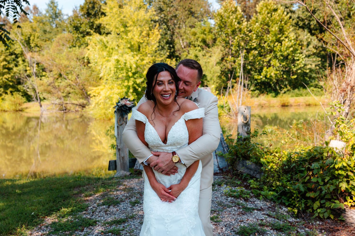 Bride and groom embrace outdoors near a pond surrounded by lush green trees on their wedding day.