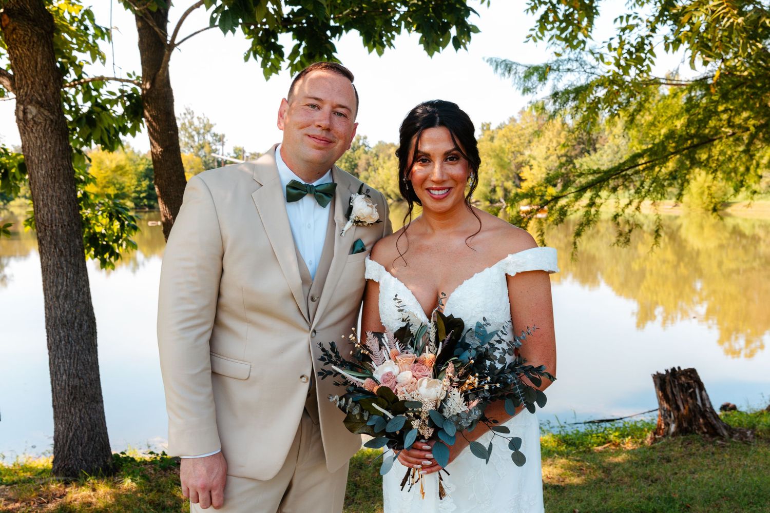 Bride and groom smiling outdoors by a lake, bride holding bouquet, groom in tan suit with bow tie.