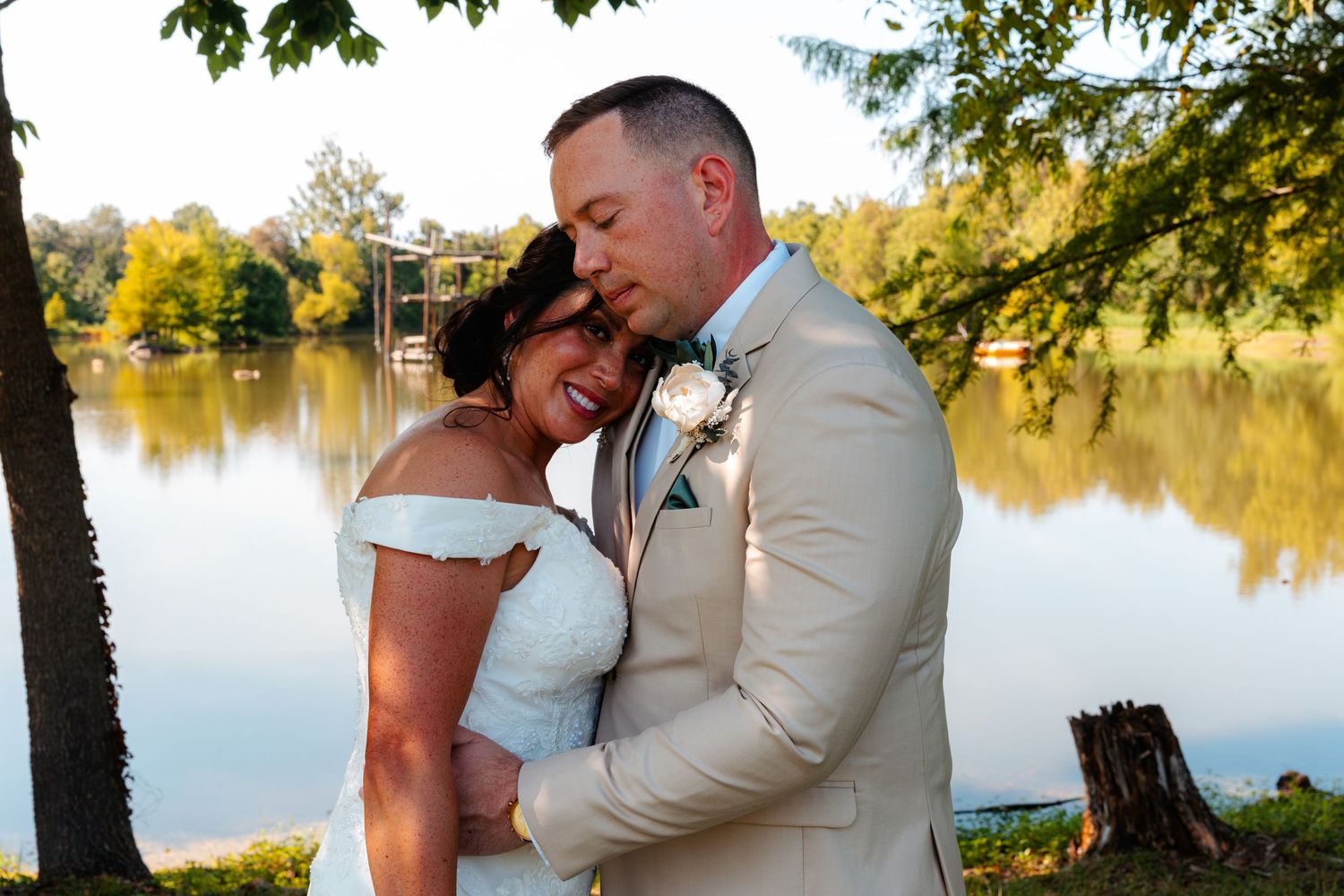 Bride and groom embrace near a serene lake surrounded by autumn trees on their wedding day.