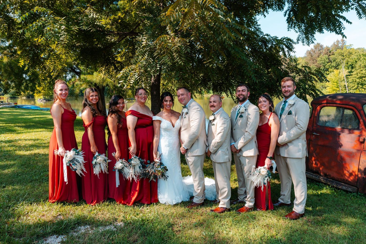 Wedding party in red dresses and tan suits posing outdoors under trees near a vintage red truck.