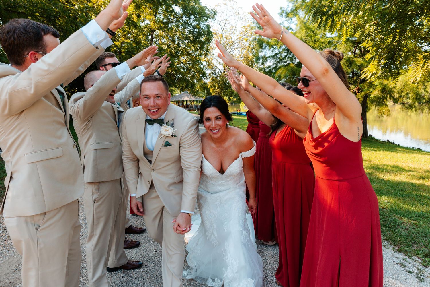 Bride and groom walk through cheering wedding party in red dresses and tan suits outdoors near a lake.
