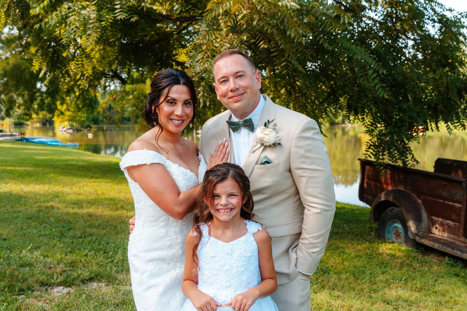 A bride and groom pose with a young girl in a white dress outdoors at a wedding reception near a pond.