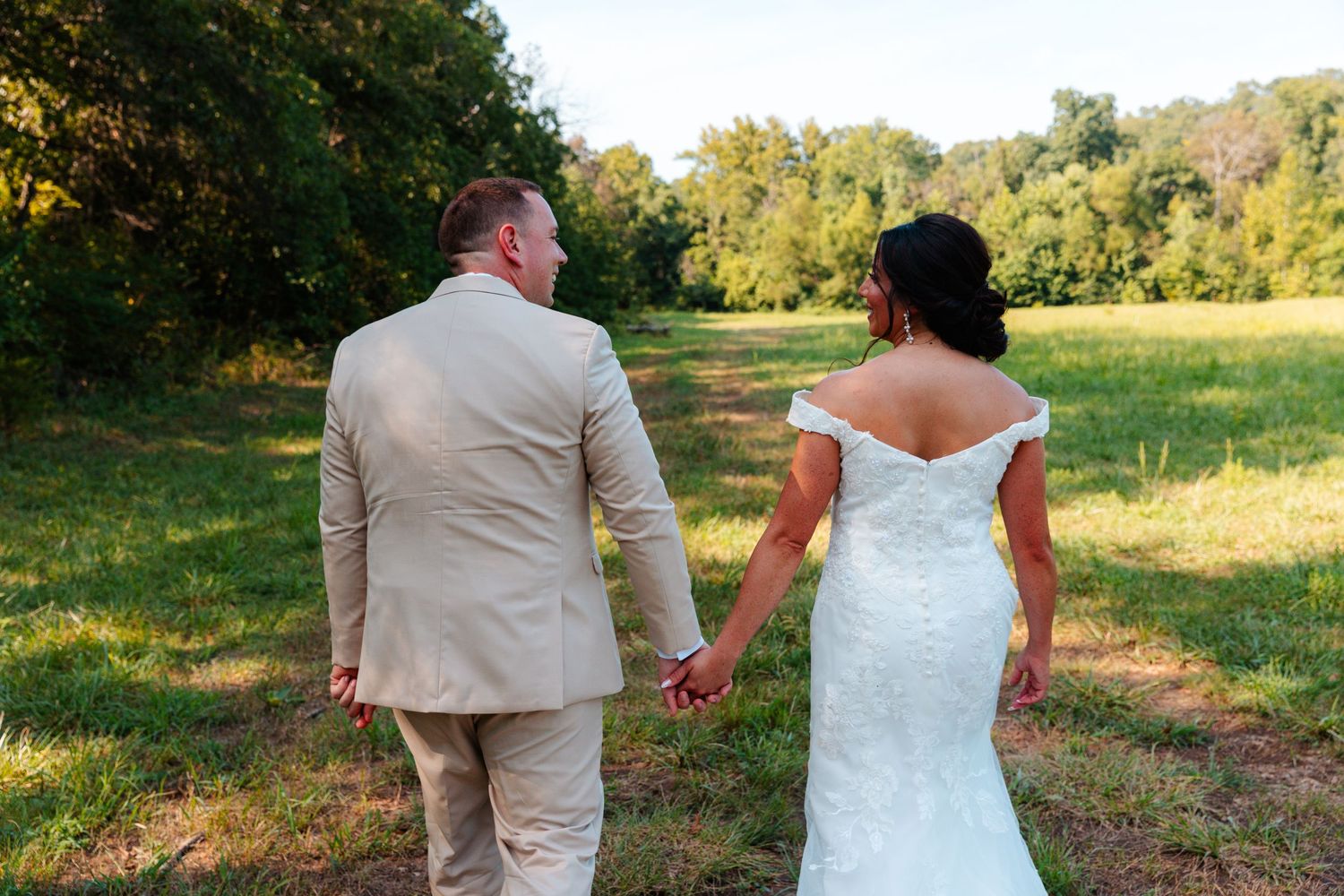 Bride and groom holding hands walking through a lush green field on their wedding day.