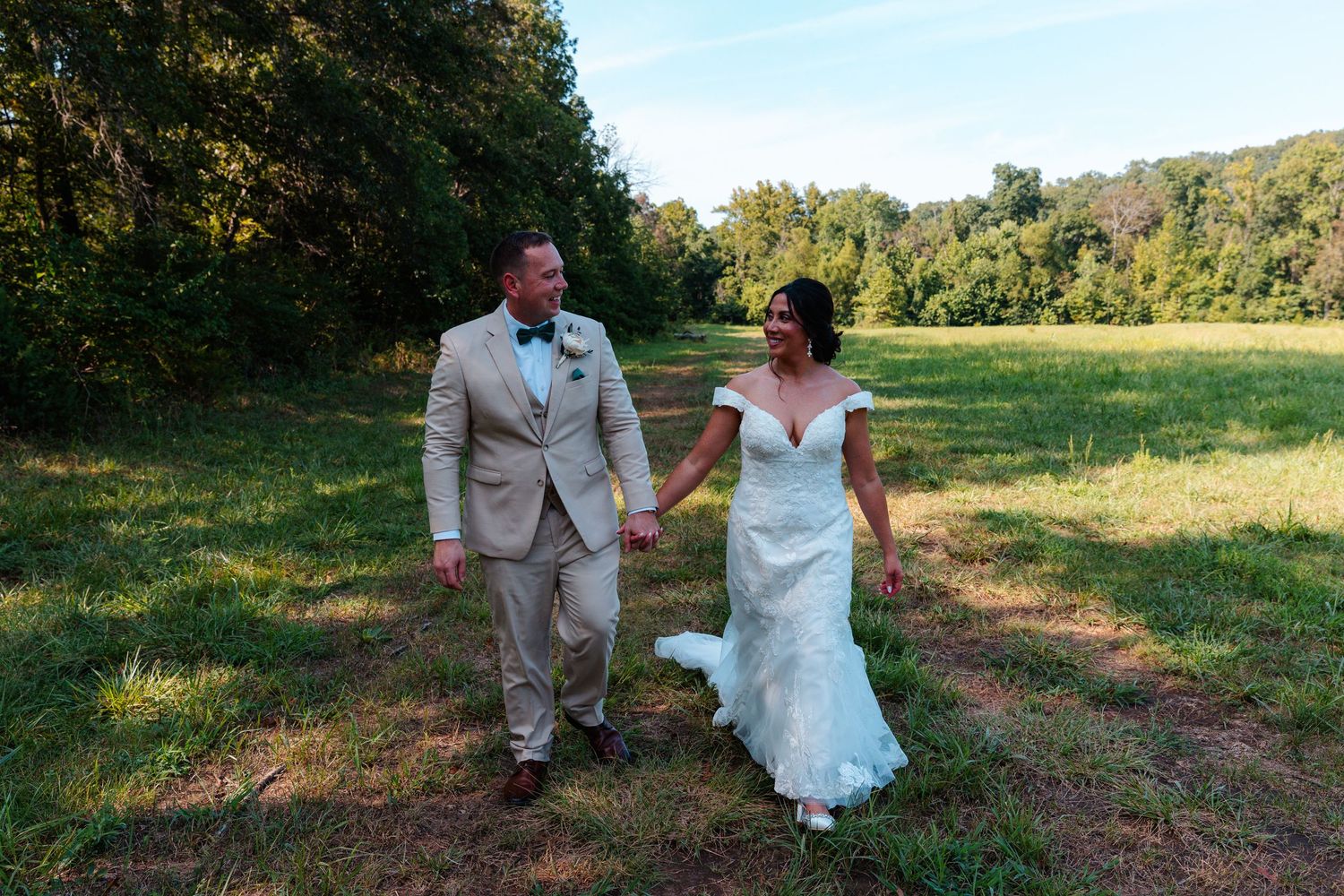 Bride and groom holding hands walking through a sunlit green meadow on their wedding day.