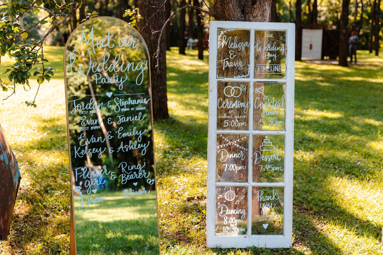 Elegant wedding seating chart displayed on a mirror and vintage window frame with white calligraphy, set outdoors on green grass.