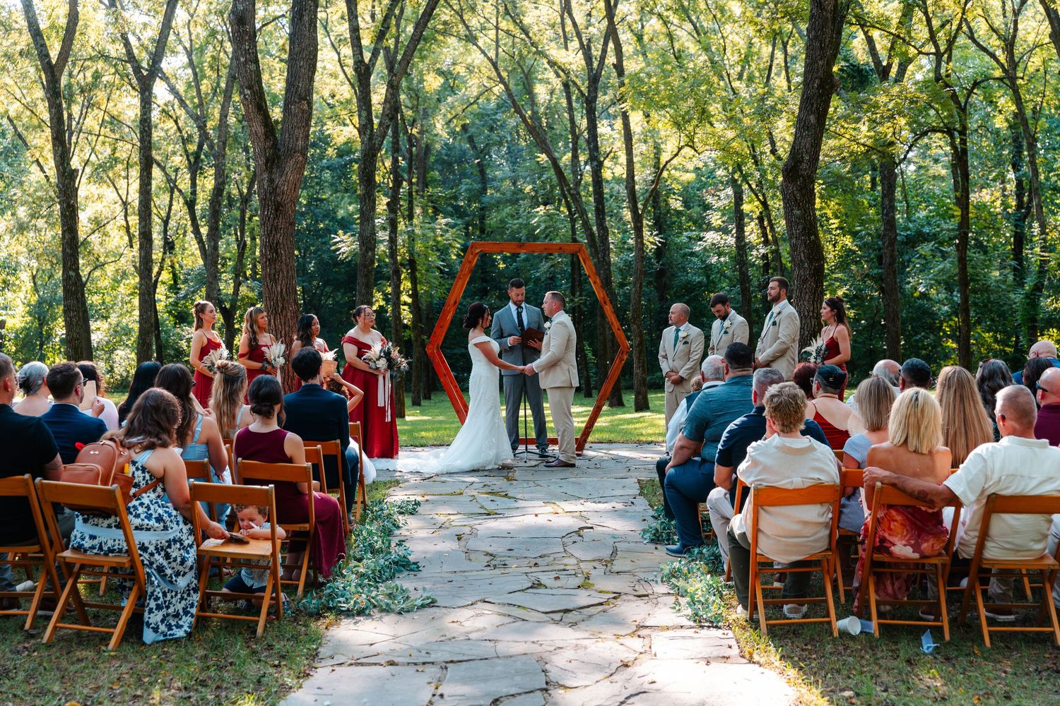 Outdoor wedding ceremony in a wooded setting with a hexagonal arch, stone aisle, and guests seated on wooden chairs.