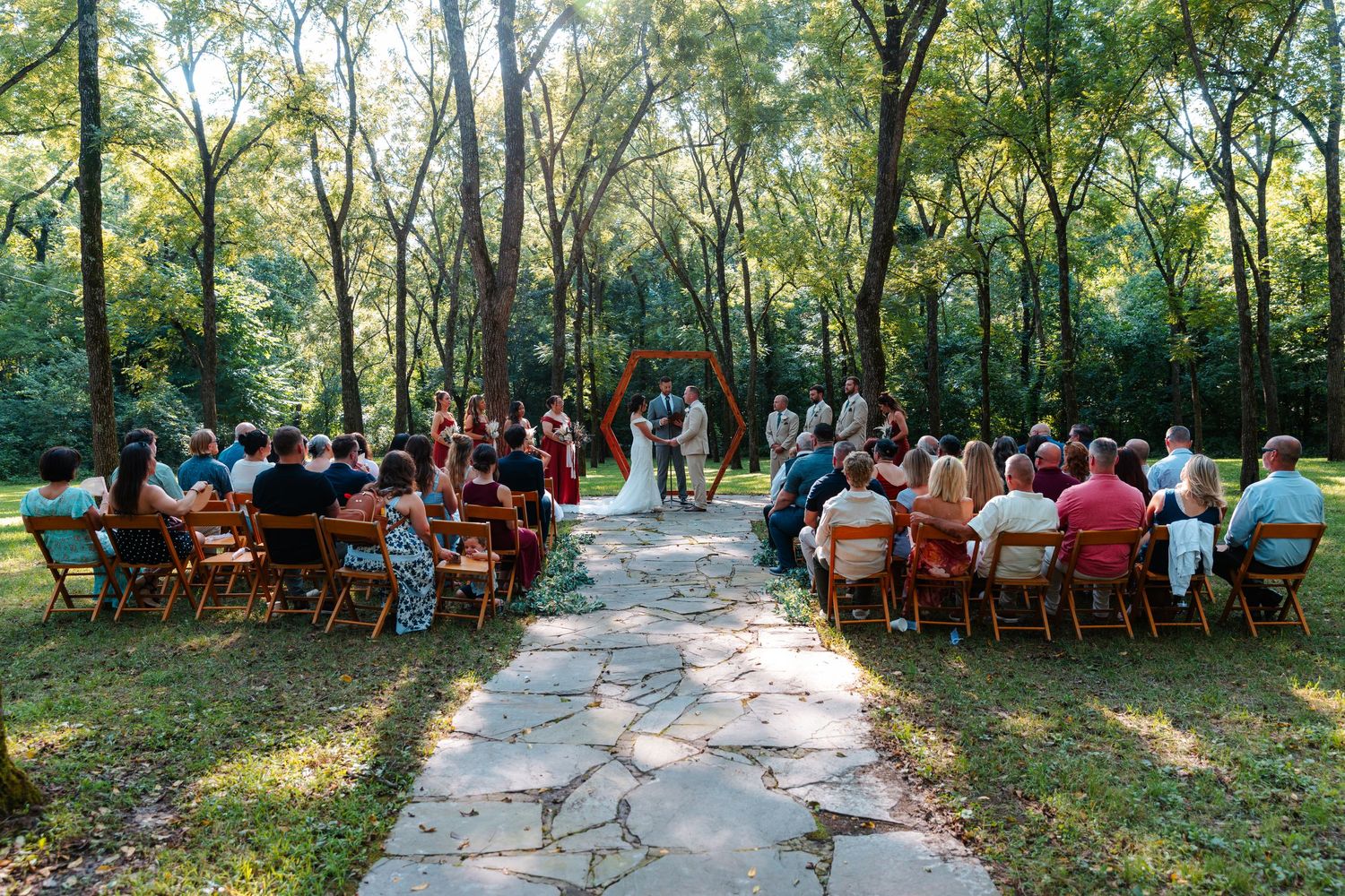 Outdoor woodland wedding ceremony with guests seated on wooden chairs along a stone path leading to a floral arch.