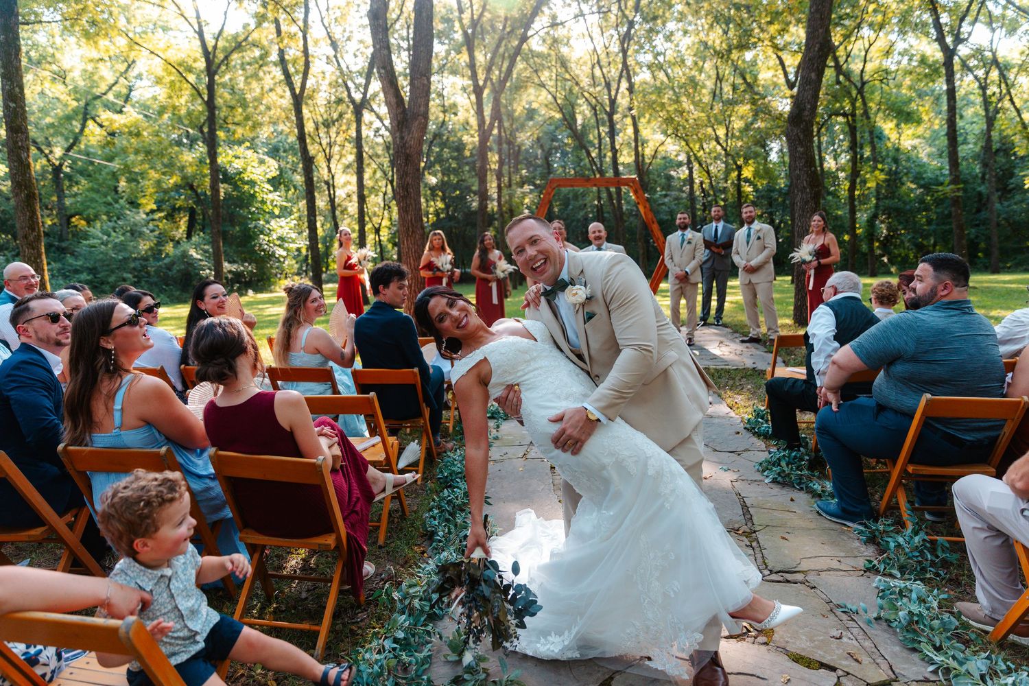 Groom dips bride during outdoor woodland wedding ceremony while guests cheer along stone aisle.
