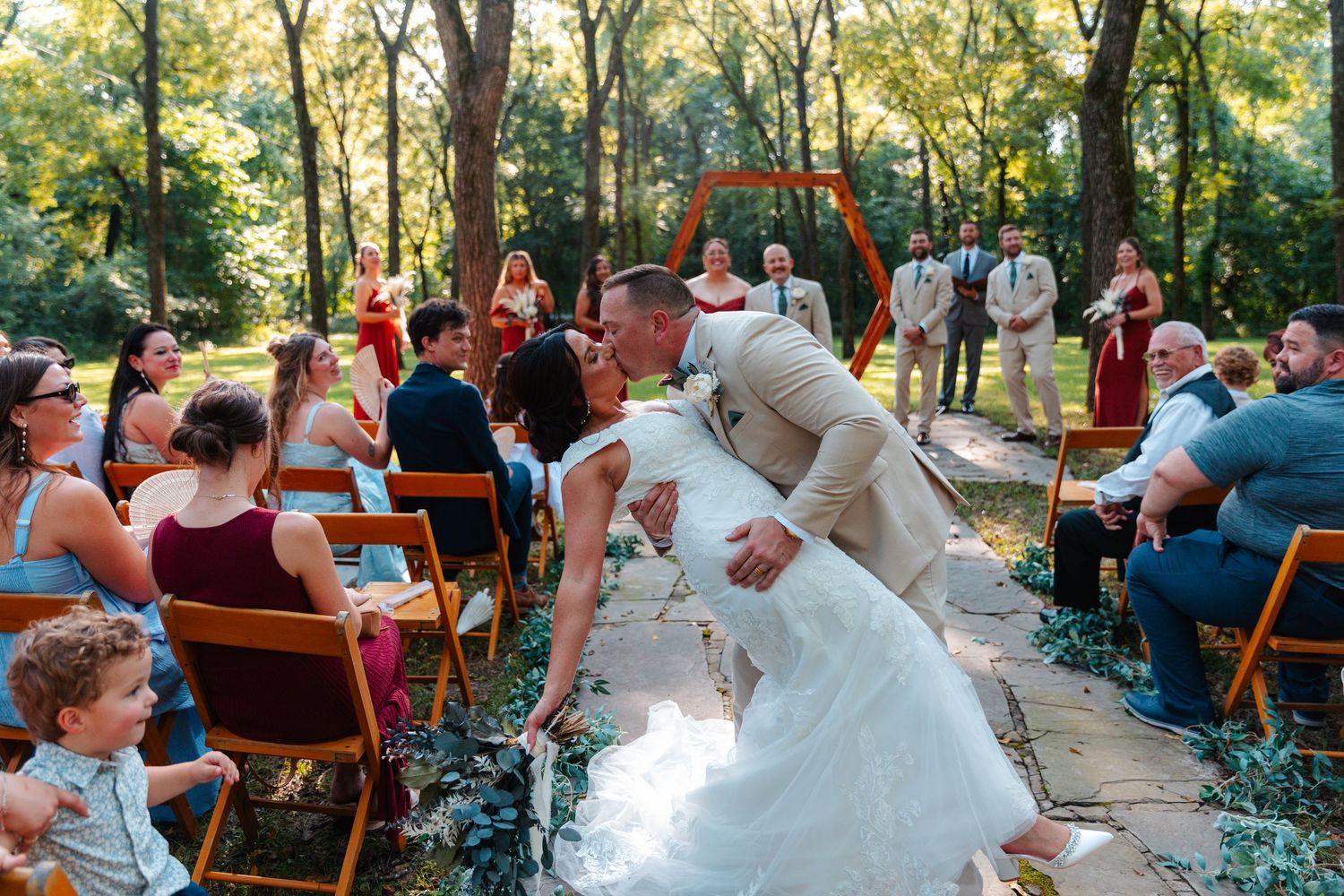 Couple shares romantic dip kiss at outdoor forest wedding ceremony with seated guests watching.