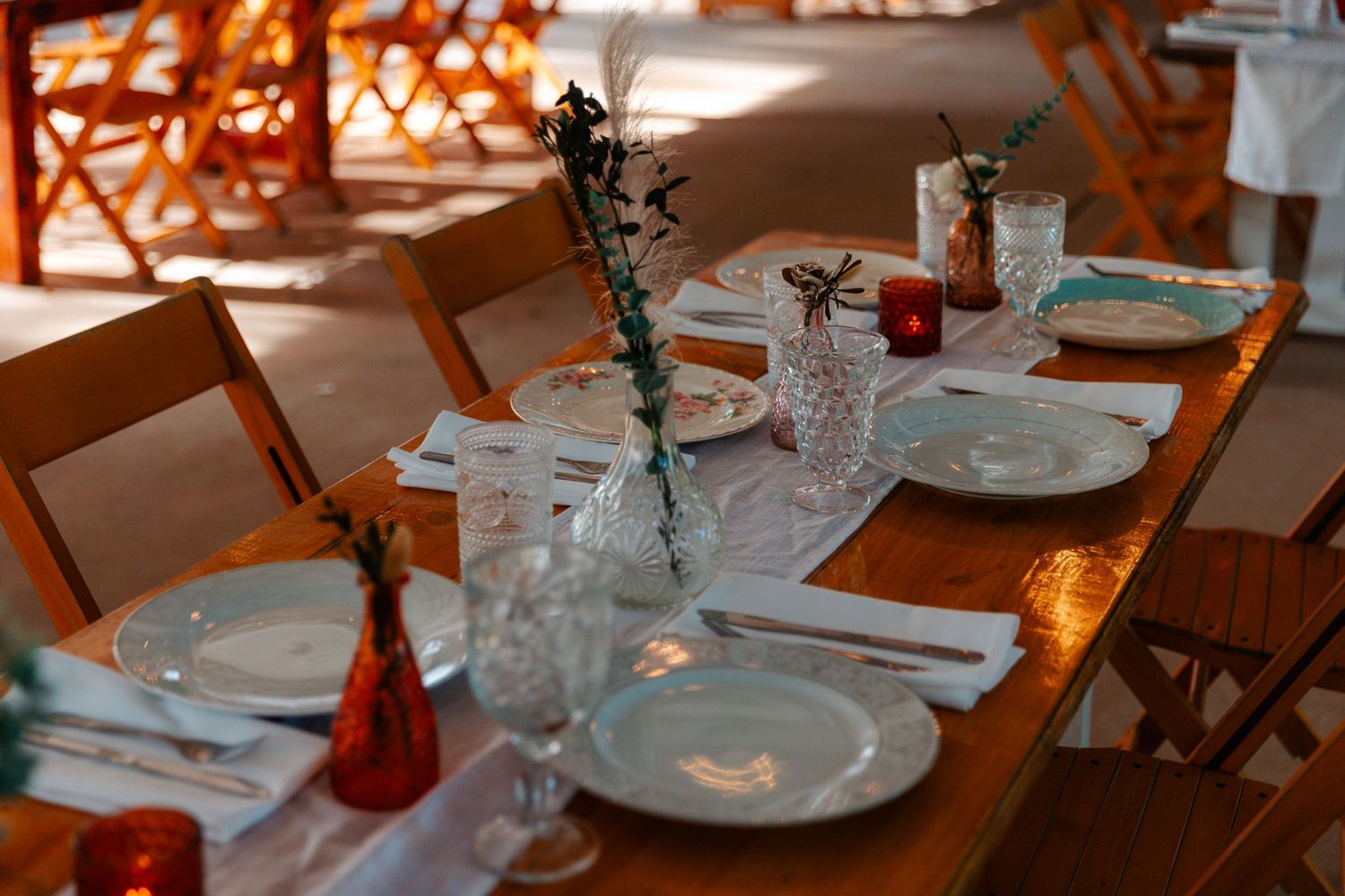 Elegantly set wooden dining table with white plates, crystal glasses, and floral bud vases in a warm outdoor setting.