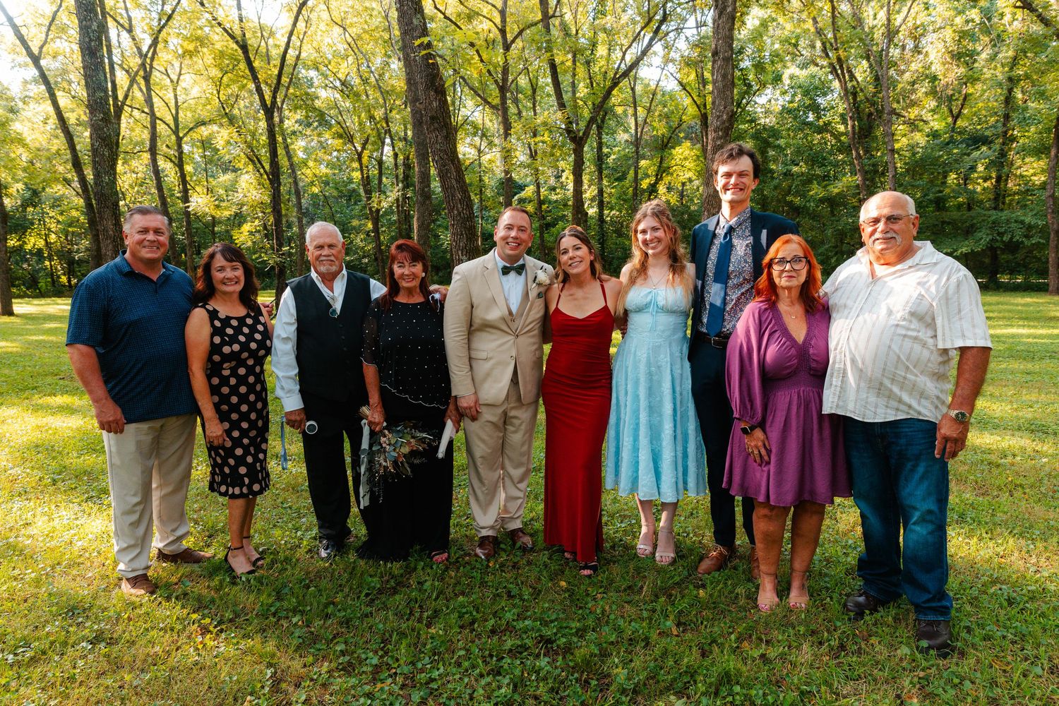 Group of ten adults posing together outdoors on green grass surrounded by trees at a formal gathering.