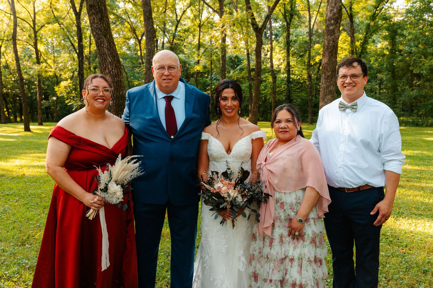Wedding party poses outdoors in wooded setting, bride in white gown with bouquet flanked by family in formal attire.
