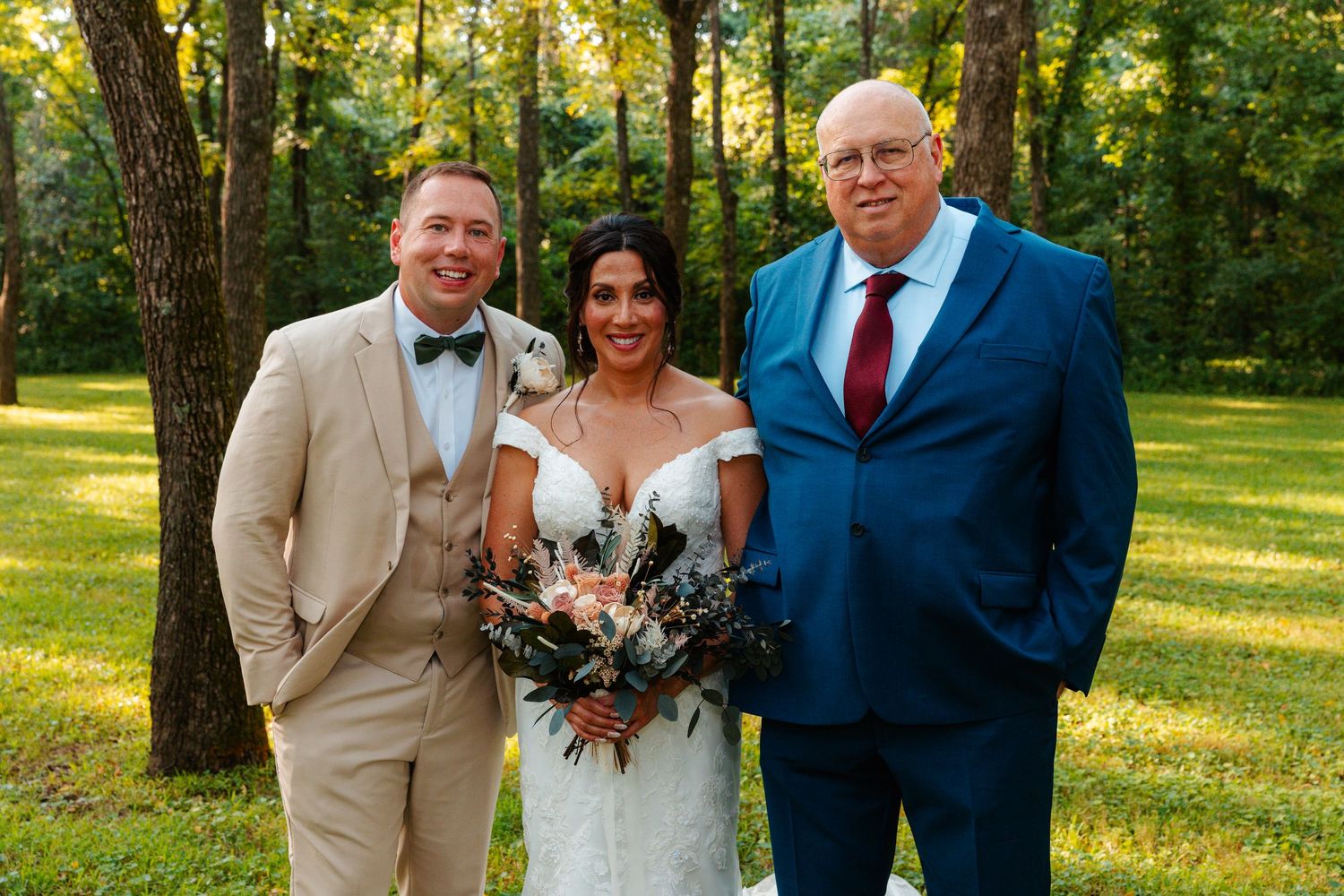 Bride in white gown holding bouquet stands with two men in suits outdoors at a wooded wedding venue.