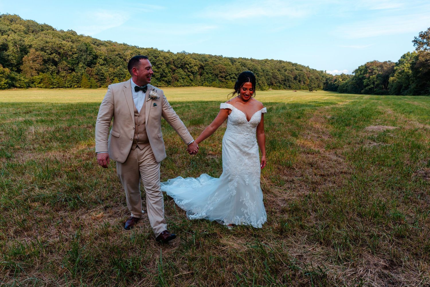 Bride and groom holding hands walking through green field on wedding day, lush hills in background.