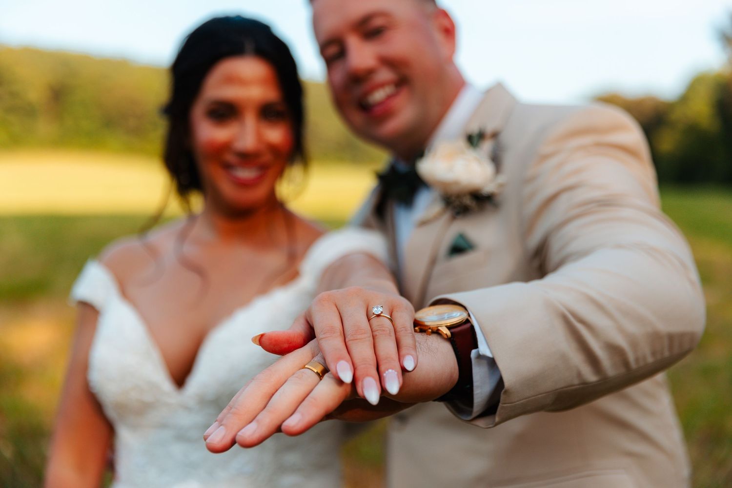 Newlywed couple smiling and showing off wedding rings outdoors in a sunny field, bride in white gown, groom in tan suit.