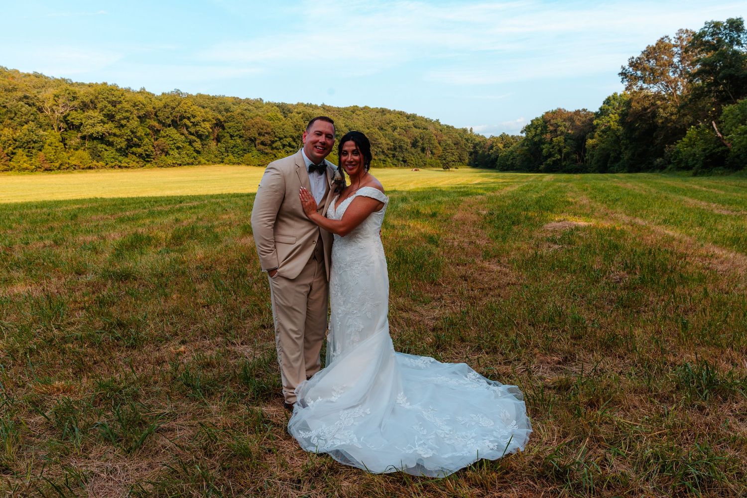 Happy bride and groom pose together in a scenic green field with rolling hills during their outdoor wedding celebration.
