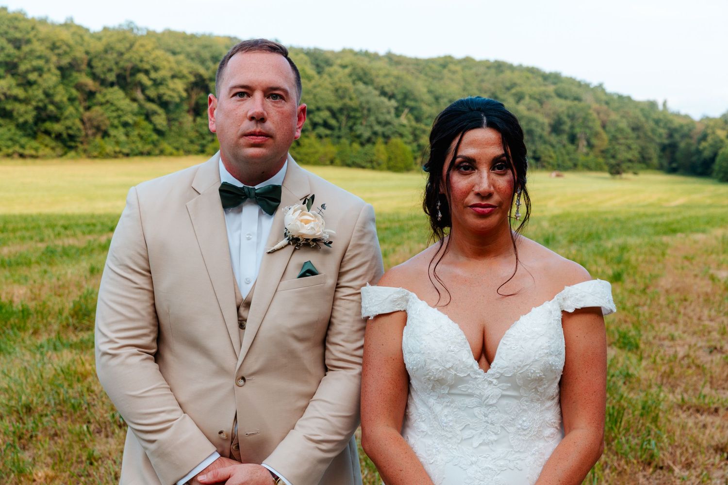 A bride and groom pose in a field, wearing wedding attire, with lush green hills in the background.
