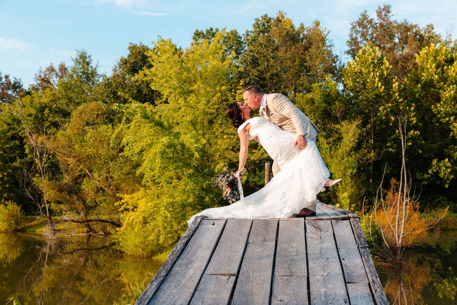 A bride and groom share a romantic dip kiss on a wooden dock surrounded by lush green trees near a serene lake.