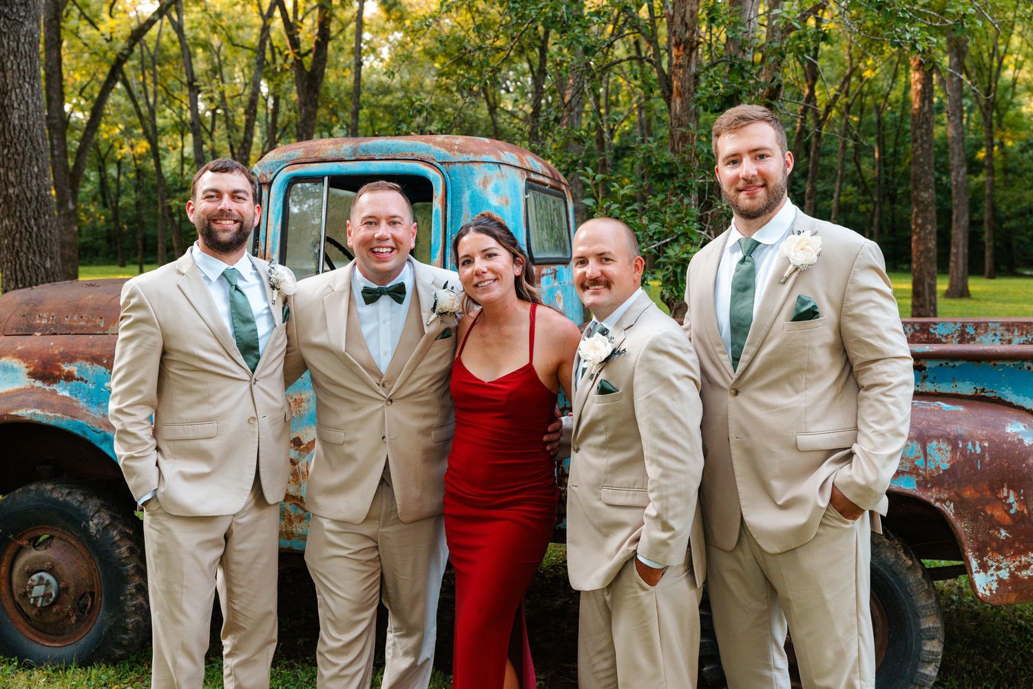 A bride in red dress poses with four groomsmen in tan suits near a rustic blue truck outdoors.