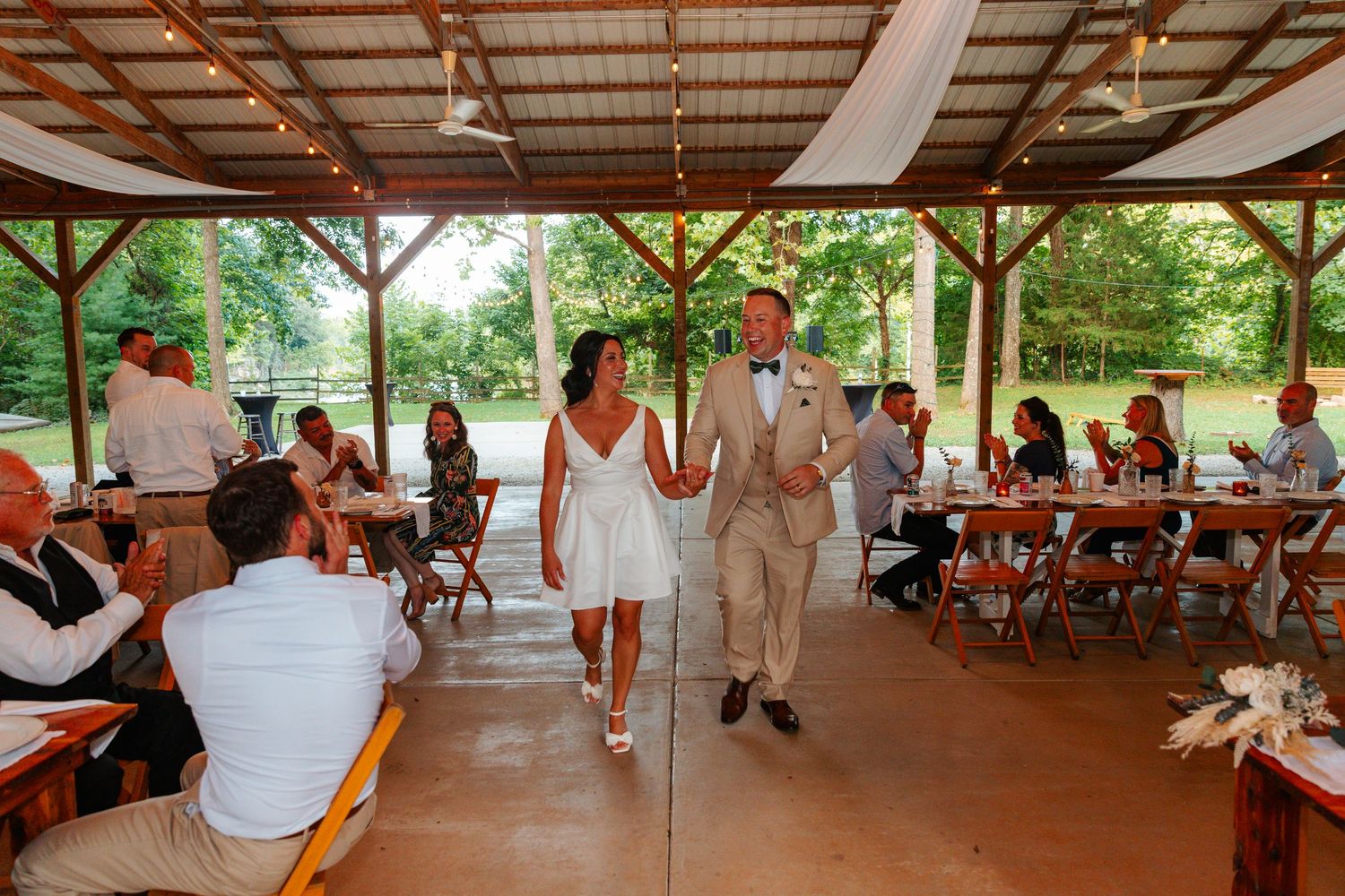 Bride and groom walk hand-in-hand through rustic outdoor wedding pavilion surrounded by seated guests.