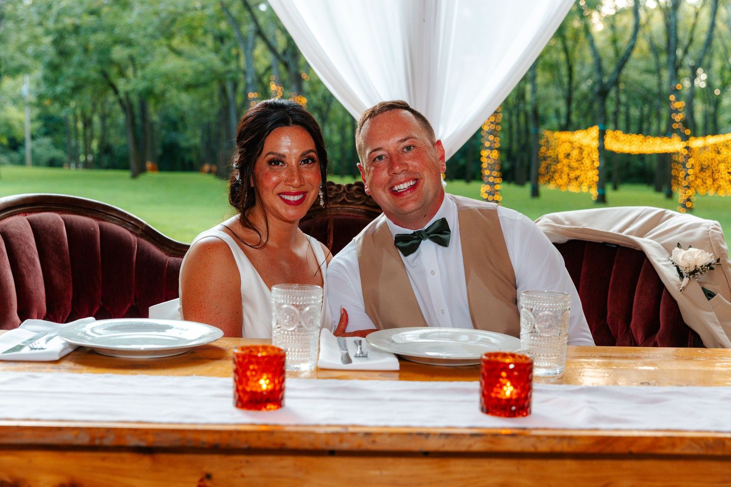 Smiling bride and groom seated at decorated wedding reception table with red candles and lush green outdoor backdrop.