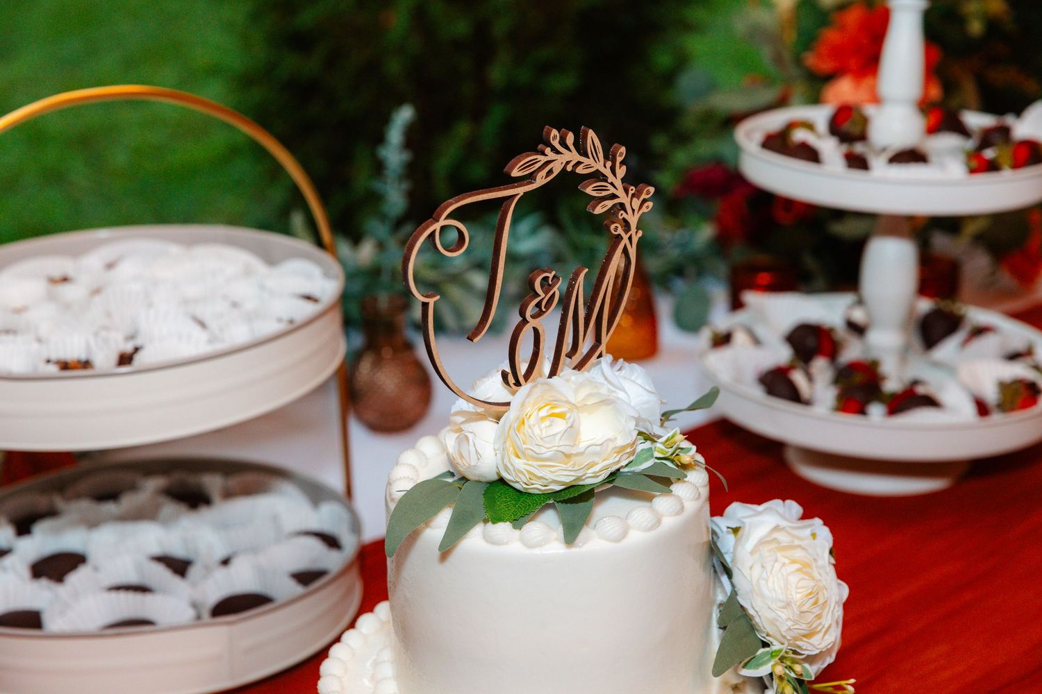 White wedding cake with floral topper and monogram, surrounded by dessert displays at an outdoor reception.