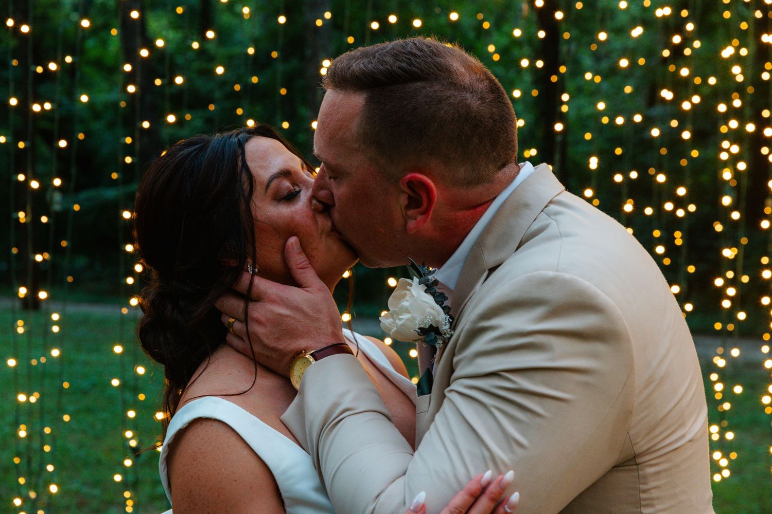 Bride and groom share first kiss at romantic outdoor wedding with twinkling fairy lights backdrop.