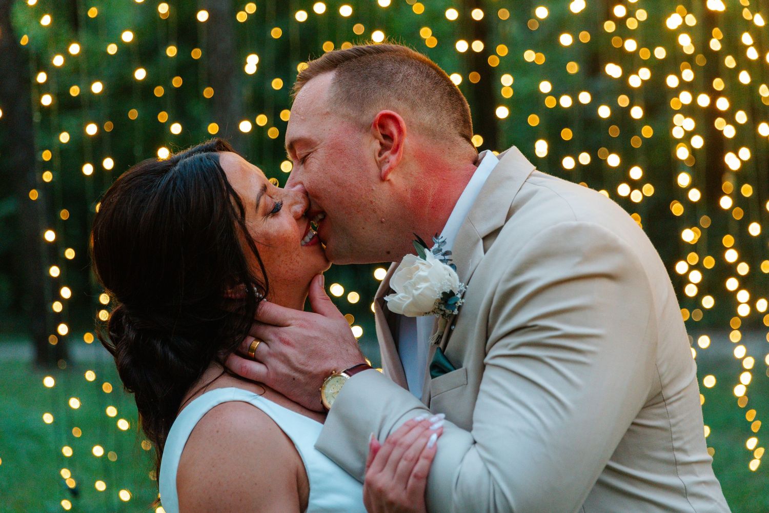 Bride and groom share first kiss at outdoor wedding with bokeh string lights glowing in background.