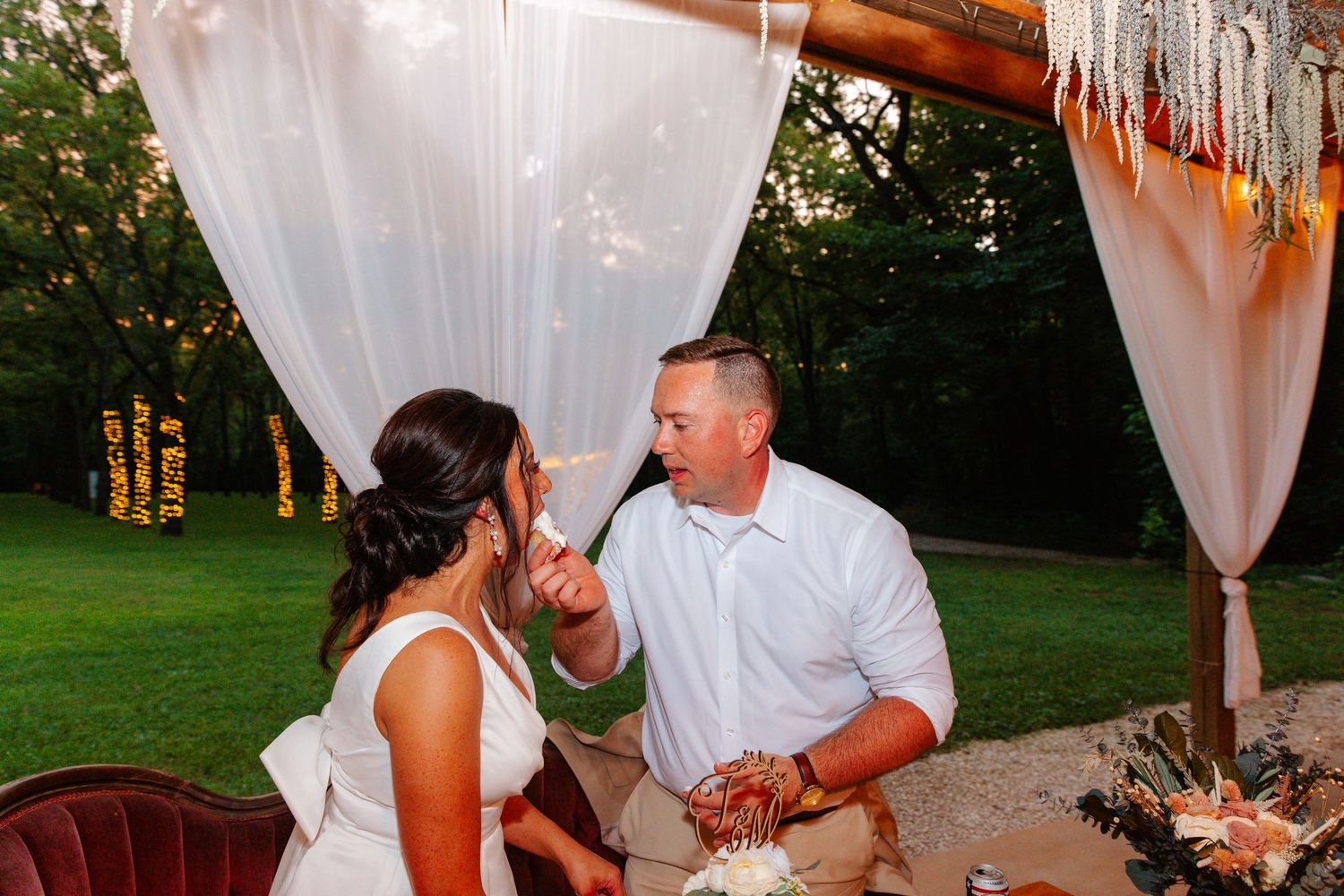 Bride and groom share a tender cake-feeding moment under a boho draped canopy at an outdoor wedding reception.