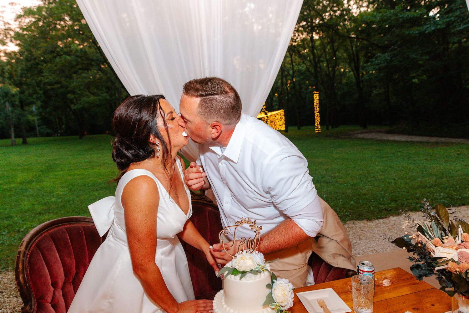 Bride and groom sharing a kiss at their wedding reception table with a white cake in a lush outdoor setting.