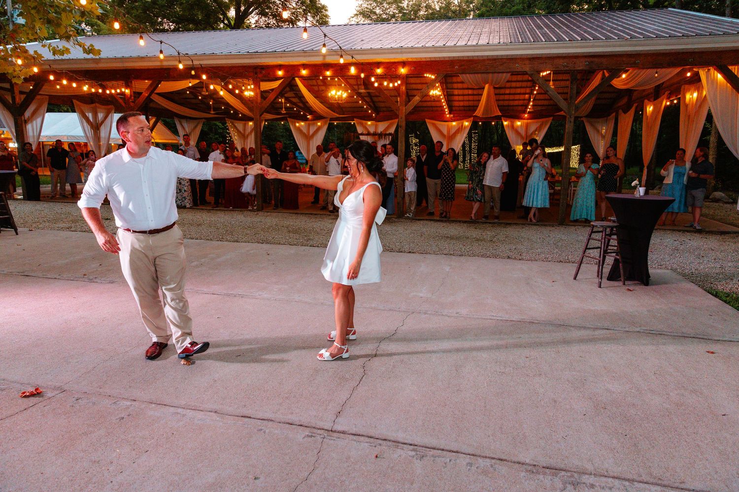 Couple dancing outdoors at a rustic wedding reception venue with string lights and guests in the background.