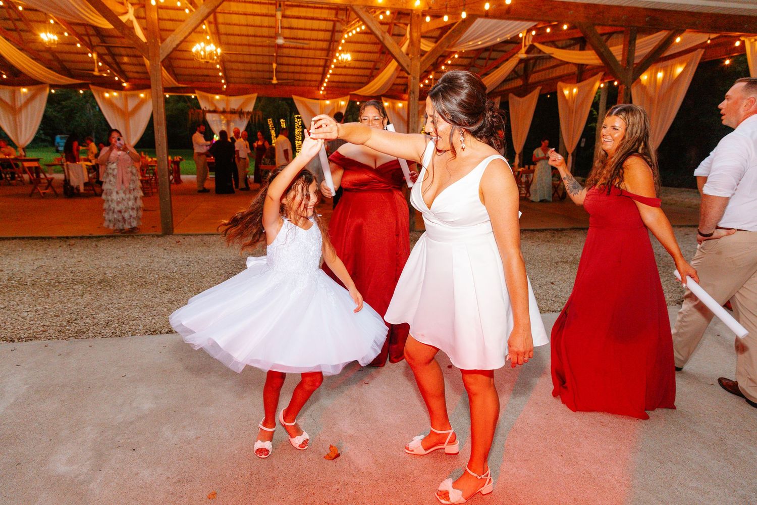 Women in red and white dresses dancing joyfully at a rustic wedding reception with string lights.