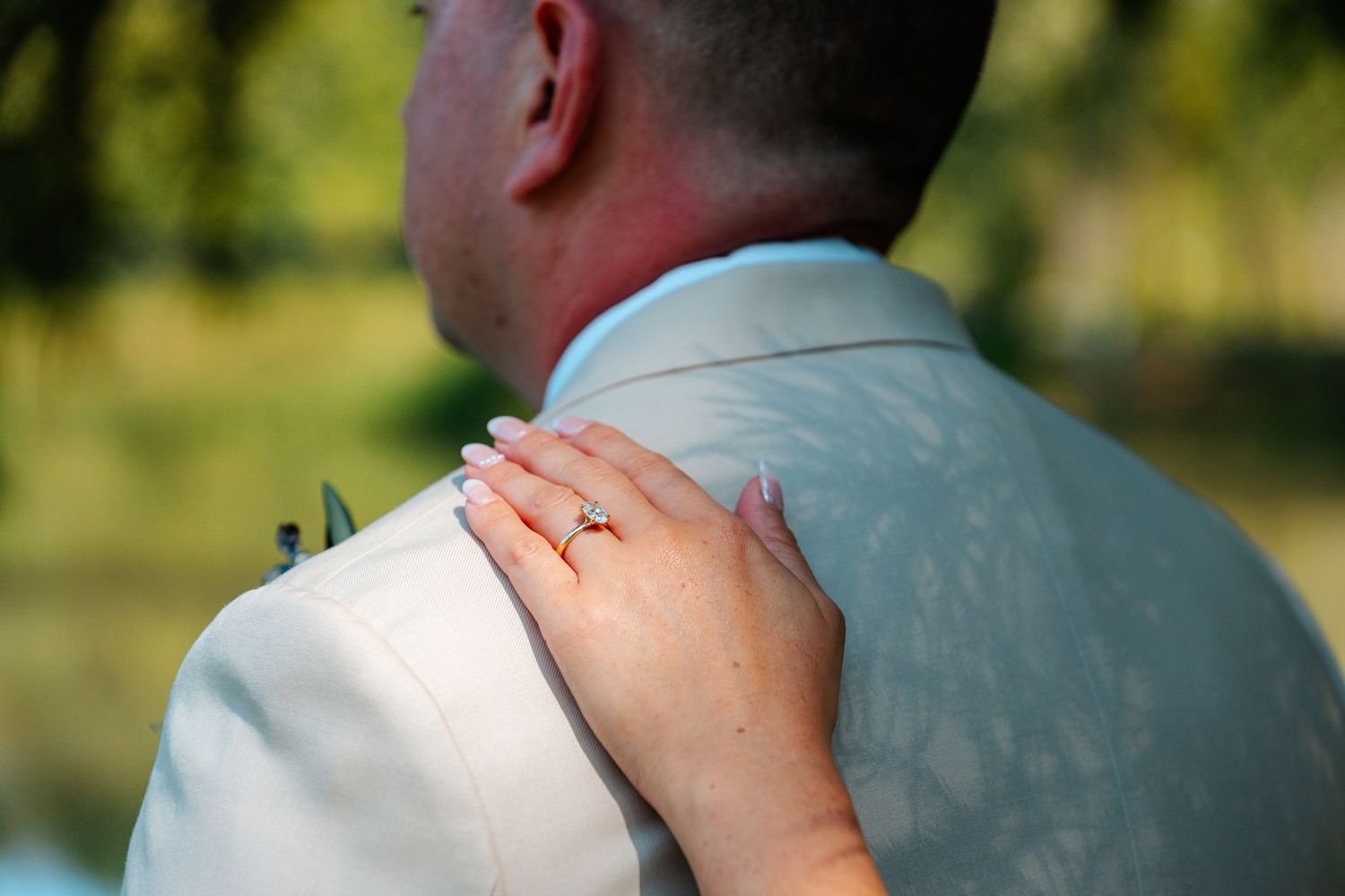 A woman's hand with an engagement ring rests on a man's shoulder in a romantic outdoor wedding moment.