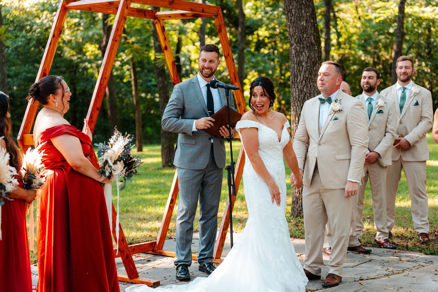 Bride and groom exchange vows outdoors under a wooden arch, with bridesmaids in red and groomsmen in tan suits.