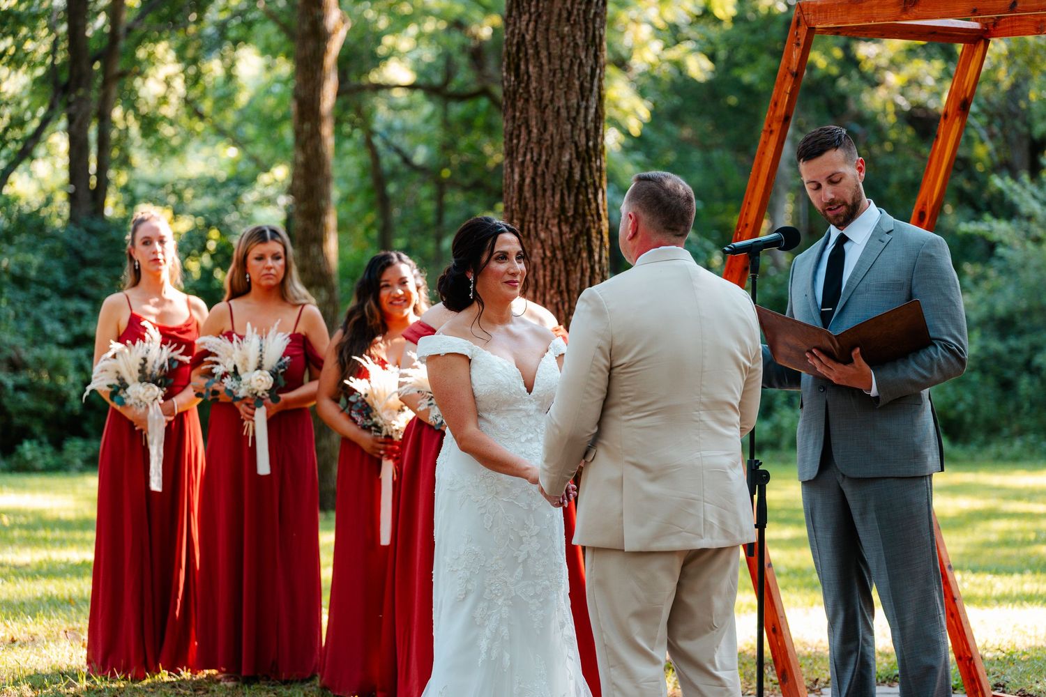 Outdoor wedding ceremony with bride and groom holding hands, bridesmaids in red dresses, and officiant reading vows under trees.