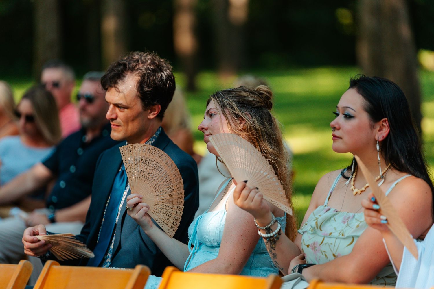 Three people sitting outdoors fanning themselves with hand fans at a summer outdoor event in a park.