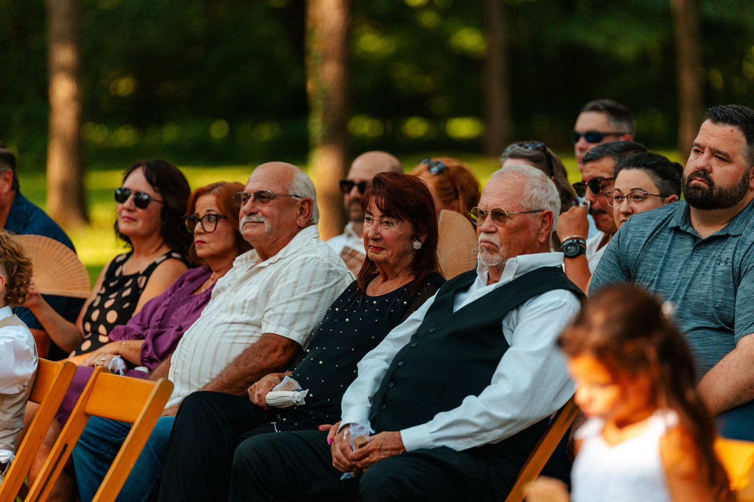 Guests seated outdoors at a wedding ceremony, enjoying the event surrounded by lush green trees in warm evening light.