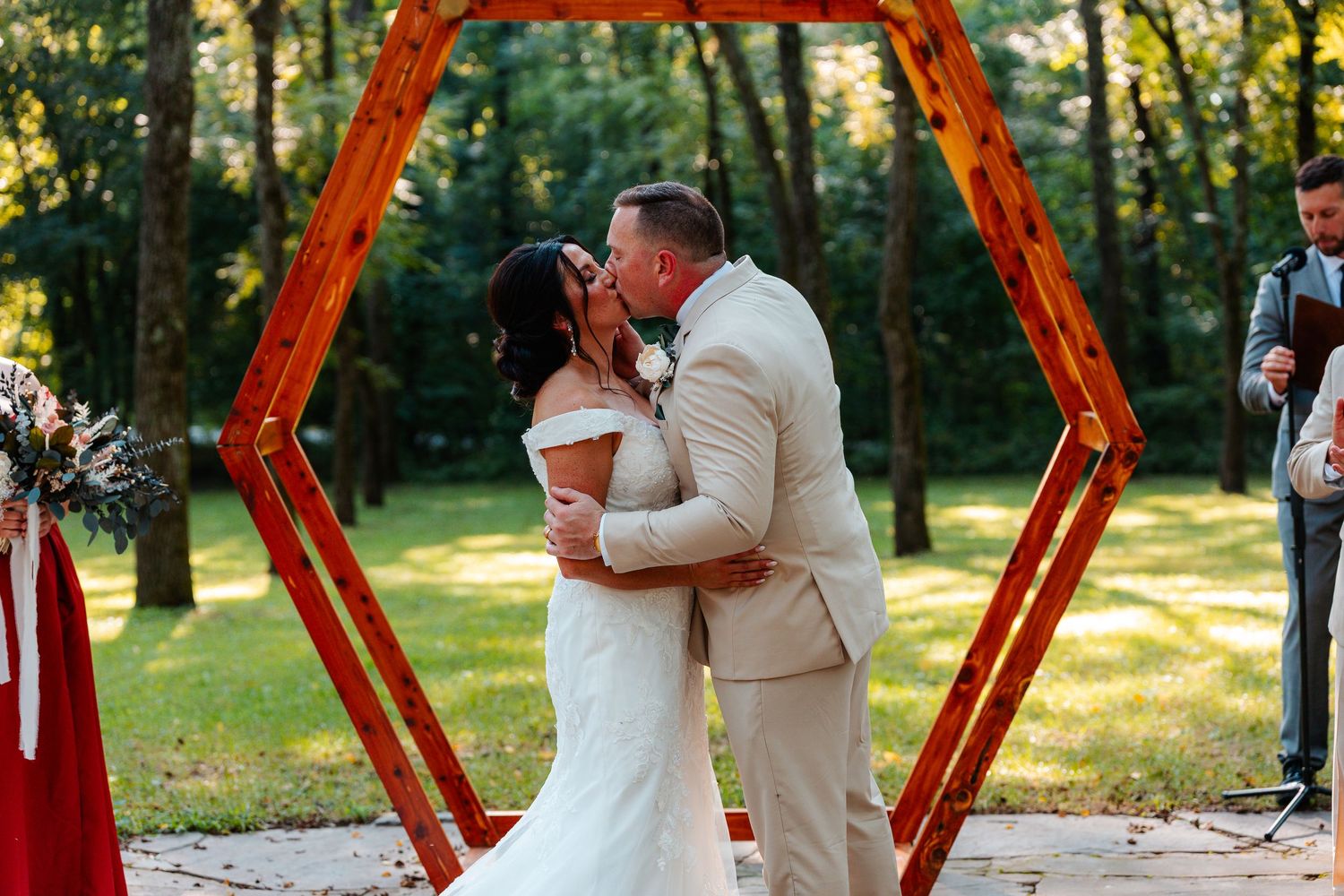 Bride and groom share first kiss under rustic wooden hexagon arch outdoors at elegant garden wedding ceremony.